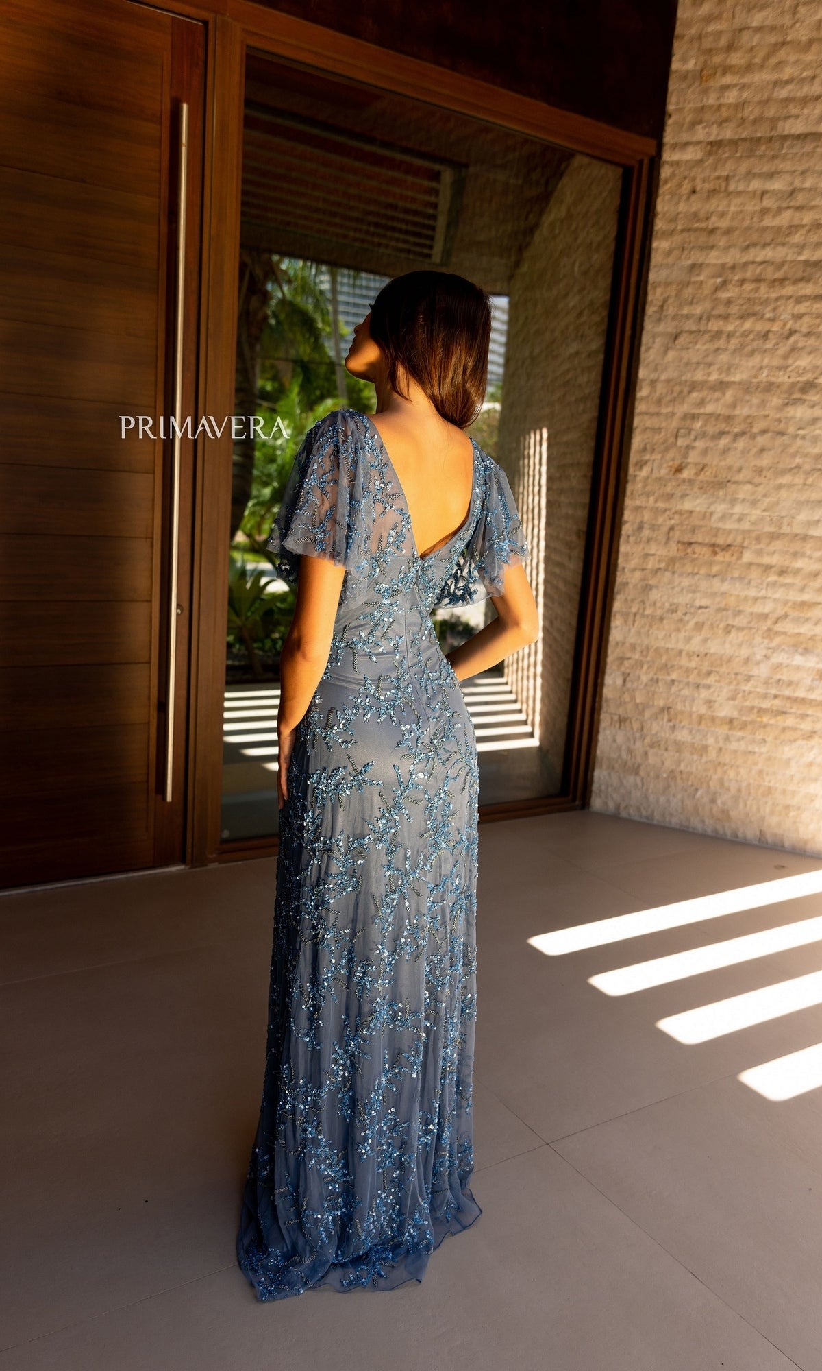 A woman in the Cape-Sleeve Beaded Formal Dress: Primavera 13124 stands facing a wooden door, natural light casting shadows across the floor.